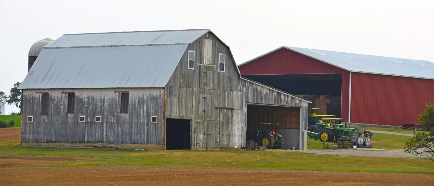 two barns at the edge of a field with tractors parked