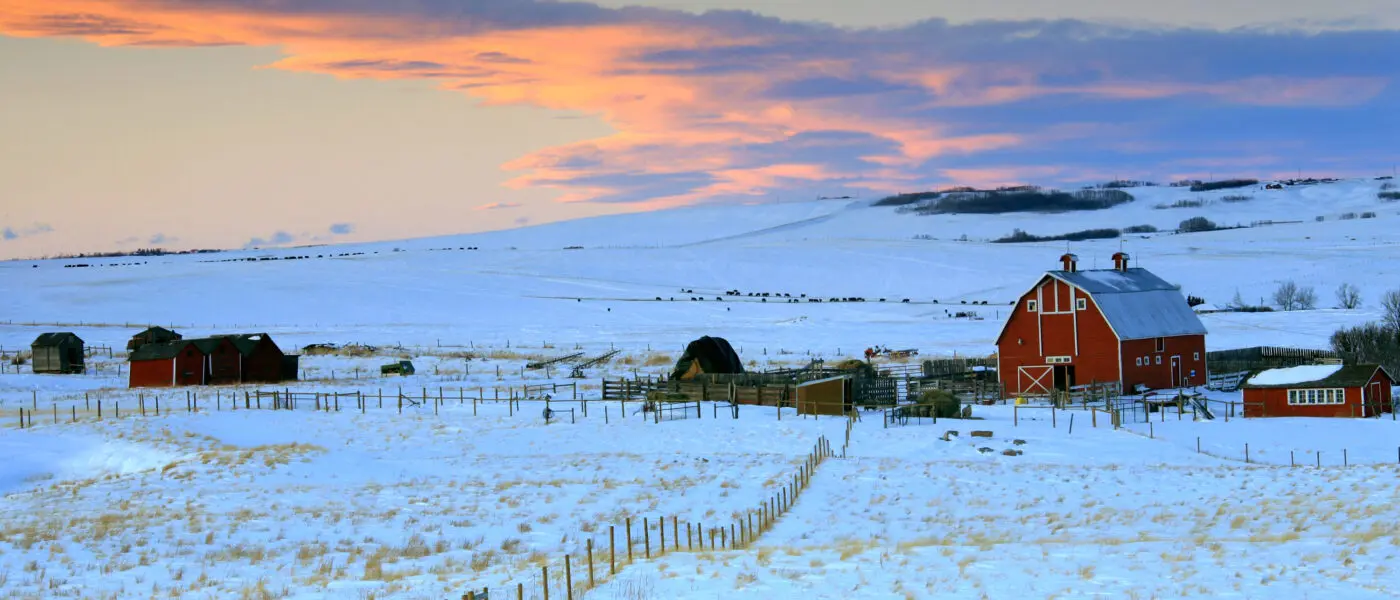 Snow-covered farm with red barn at sunset.