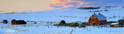 Snow-covered farm with red barn at sunset.