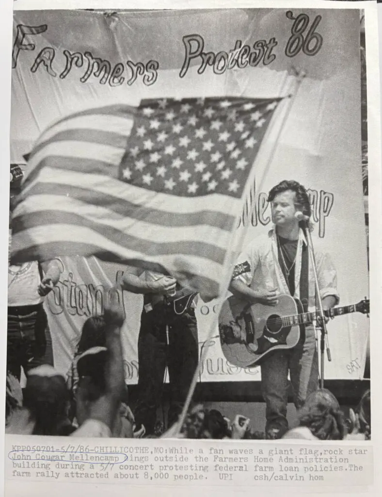 John Mellencamp performing in front of a Farmers Protest '86 flag