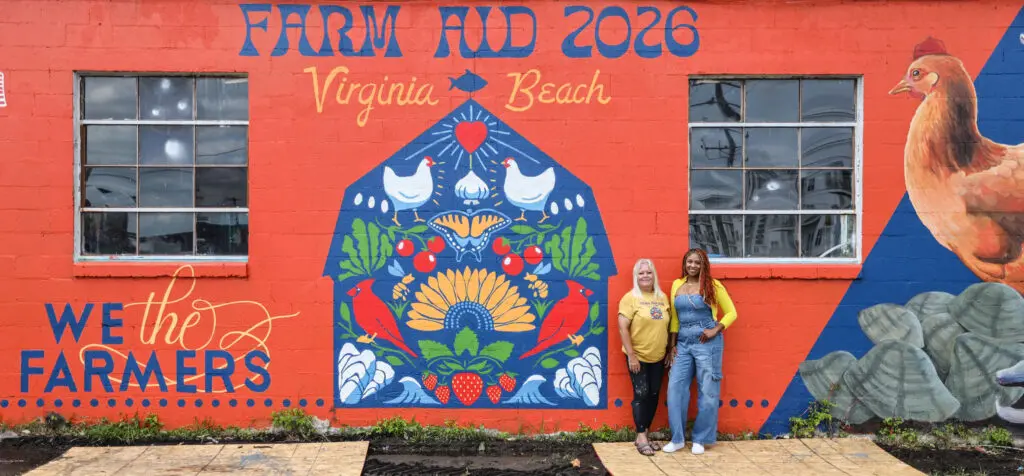 Farm Aid mural with the artists. Mural has a barn with Farm Aid 2026, Virginia Beach, a chicken, oysters, "We the Farmers"