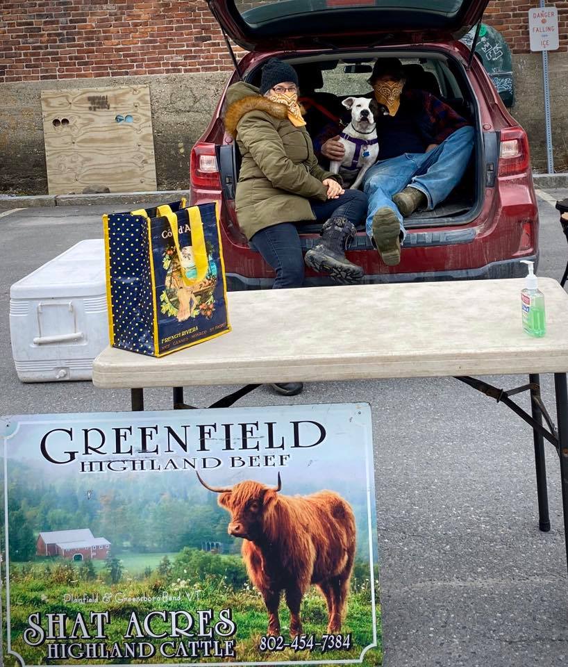 Two people wearing masks and winter coats sit in an open car trunk in a parking lot, behind a folding table set with packaged beef and hand sanitizer.