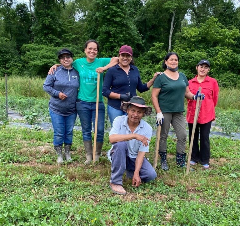 A group of Latino farmers stands, with one farmer kneeling, with farming tools in hand.