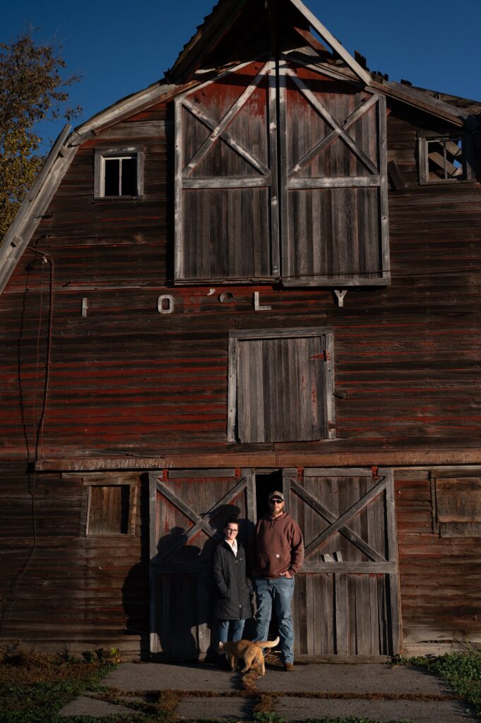 two people stand in front of a barn with a cat at their feet